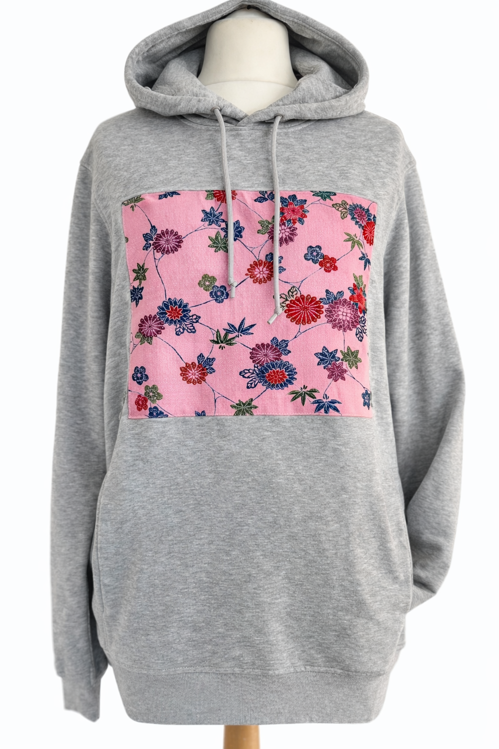 Grey pullover hoodie with pink Japanese floral textile panel, handmade statement hoodie combining modern streetwear and traditional Japanese fabric, shown on mannequin against white background.