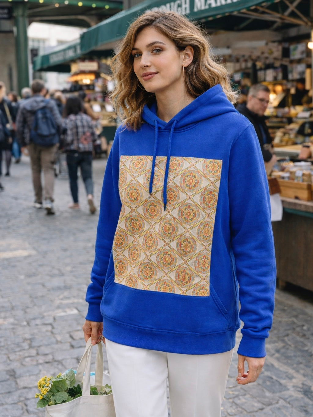 Royal blue hoodie with side pockets and a large vintage kimono obi silk panel on the front, worn by a European woman at an upscale outdoor market, styled with tailored white trousers and a neutral tote bag, natural light highlighting the geometric floral pattern and creating a refined, contemporary look.