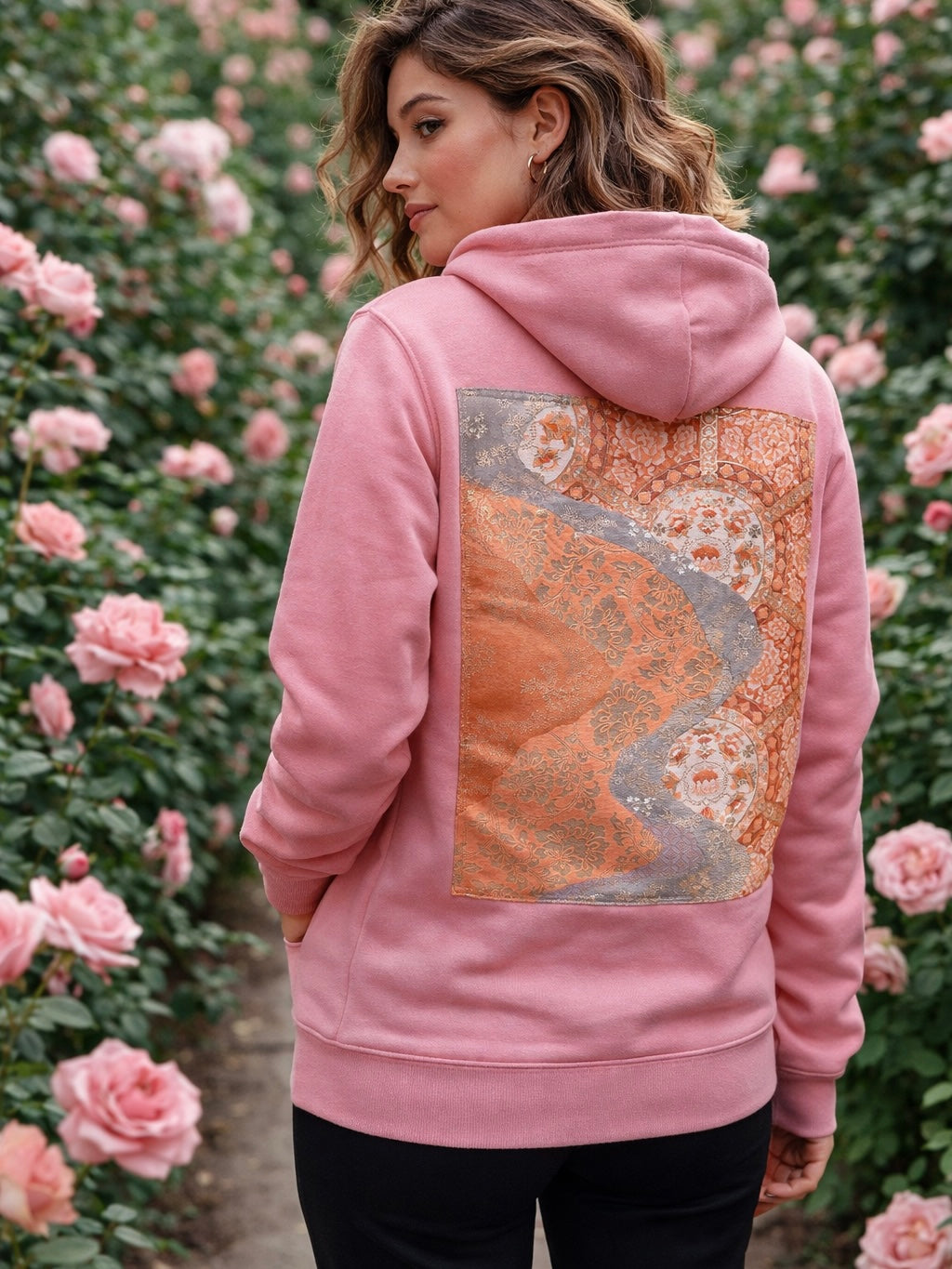 Pink kimono obi hoodie in size S worn by a European woman in a blooming rose garden, shown from the back to highlight the large coral and gold vintage obi silk panel with intricate floral and ornamental patterns, fitted silhouette with a neat shoulder line and slightly tailored shape through the waist, soft natural light enhancing the romantic garden setting.