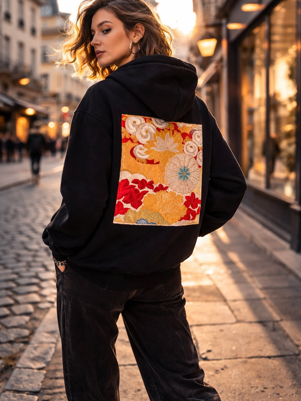 Black unisex hoodie with a vintage Japanese obi panel stitched on the back in gold, red and cream floral pattern, shown on a female model in an urban European street setting.